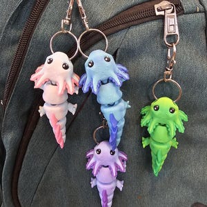 Op de afbeelding: Een verzameling kleurrijke axolotl sleutelhangers in roze, blauw, paars en groen. Elke sleutelhanger heeft een gedetailleerd, driedimensionaal axolotl ontwerp met een ring en sluiting. De sleutelhangers zijn bevestigd aan een teal rugzak.