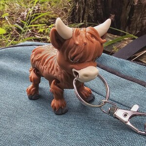 Op de afbeelding: Een bruin en bruin Highland koeienbeeldje met witte hoorns en een zilveren sleutelhanger. De koe heeft een ruige vacht en staat op een blauw oppervlak. De achtergrond omvat groen gebladerte en een boomstam.