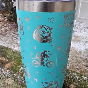 May include: A turquoise stainless steel tumbler with a silver lid. The tumbler has a repeating pattern of opossums in various poses, including reading a book, riding a bicycle, and holding a baby opossum. The opossums are outlined in silver and have a heart pattern in the background.