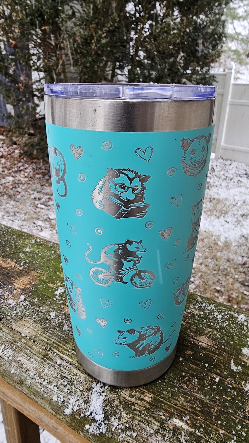 May include: A turquoise stainless steel tumbler with a silver lid. The tumbler has a repeating pattern of opossums in various poses, including reading a book, riding a bicycle, and holding a baby opossum. The opossums are outlined in silver and have a heart pattern in the background.