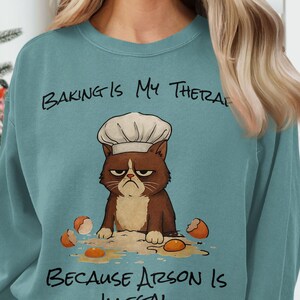 May include: Teal sweatshirt with a grumpy cat wearing a chef's hat, surrounded by broken eggs. The text reads "Baking is My Therapy Because Arson Is Illegal."