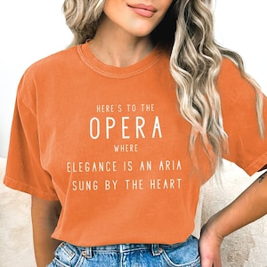May include: Orange short-sleeved t-shirt with white text that reads "Here's to the Opera where Elegance is an Aria sung by the heart."