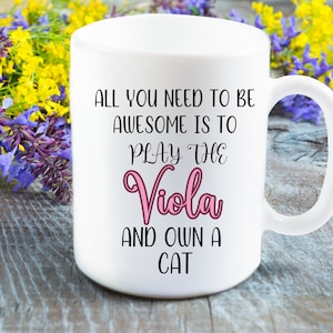 Könnte beinhalten: Weiße Keramik-Kaffeetasse mit schwarzem Text, der lautet: "Alles, was du brauchst, um großartig zu sein, ist, Viola zu spielen und eine Katze zu besitzen". Das Wort "Viola" ist in Pink.