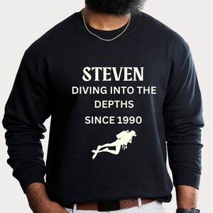 May include: Black sweatshirt with white text that reads "STEVEN DIVING INTO THE DEPTHS SINCE 1990". A white silhouette of a scuba diver is below the text.