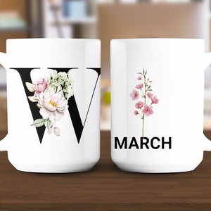 May include: Two white ceramic mugs. One mug features a large black letter "W" adorned with watercolor flowers in pink, white, and green. The other mug displays the word "MARCH" in black, with a pink floral sprig.