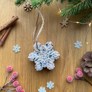 May include: A white snowflake-shaped bird feeder with a twine hanger. The feeder is filled with birdseed and is surrounded by small wooden snowflakes, cinnamon sticks, and red berries.