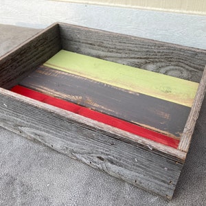 May include: A wooden tray with a rustic finish. The tray is painted in shades of gray, green, and red. The tray is perfect for displaying small items or serving snacks.