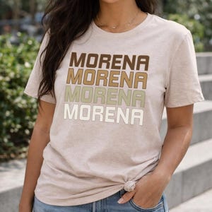 May include: Beige t-shirt with the word "MORENA" printed four times in a stacked design. The word is in shades of brown, tan, and white. The model is wearing gold hoop earrings and a gold necklace.