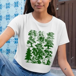 May include: White crop top with a green tree design. The graphic features various tree silhouettes in shades of green. The top is paired with light blue denim jeans.