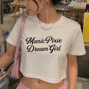 Peut inclure: Une femme porte un crop top blanc avec l'inscription noire "Manic Pixie Dream Girl" et un pantalon de survêtement rose. Elle est dans un magasin de disques, regardant des vinyles. Des lunettes de soleil roses sont sur sa tête.