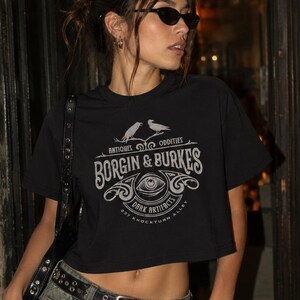 May include: A black cropped t-shirt featuring a graphic print in light gray. The design includes two birds, an eye, and the text: Antiques Oddities, Borgin & Burkes, Dark Artifacts, 237 Knockturn Alley. The shirt has short sleeves and a crew neckline.