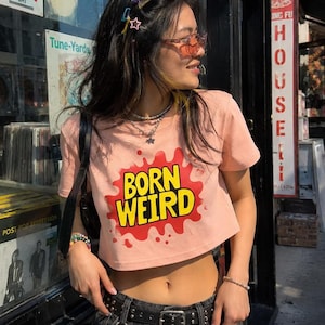 Op de afbeelding: Een perzikkleurig kort T-shirt met de tekst BORN WEIRD in gele letters op een rode achtergrond met spetters. Het shirt wordt gecombineerd met een zwarte spijkerrok, een zwarte riem met zilveren studs en een zwarte schoudertas.