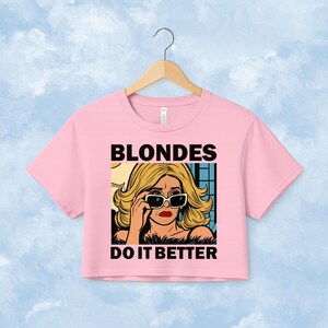 Pu&ograve; includere: Un crop top rosa chiaro con un'illustrazione grafica di una persona bionda che indossa occhiali da sole. Il testo BLONDES DO IT BETTER &egrave; stampato in grassetto nero in lettere maiuscole sopra e sotto l'illustrazione, che raffigura una persona con capelli biondi ondulati, rossetto rosso e occhiali da sole con montatura scura.