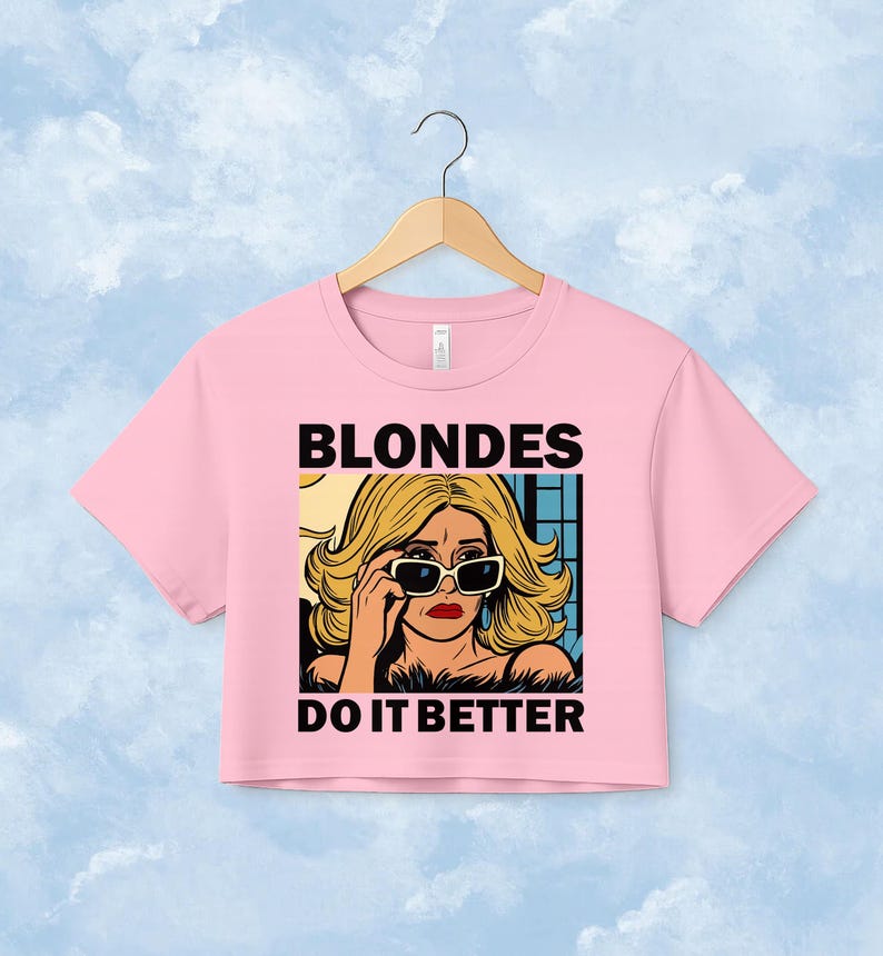 Pu&ograve; includere: Un crop top rosa chiaro con un'illustrazione grafica di una persona bionda che indossa occhiali da sole. Il testo BLONDES DO IT BETTER &egrave; stampato in grassetto nero in lettere maiuscole sopra e sotto l'illustrazione, che raffigura una persona con capelli biondi ondulati, rossetto rosso e occhiali da sole con montatura scura.