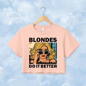 Pu&ograve; includere: Un top corto a maniche corte color pesca chiaro con un'illustrazione grafica di una persona bionda che indossa occhiali da sole. Il testo BLONDES DO IT BETTER &egrave; stampato in grassetto nero in maiuscolo sopra e sotto il disegno centrale.