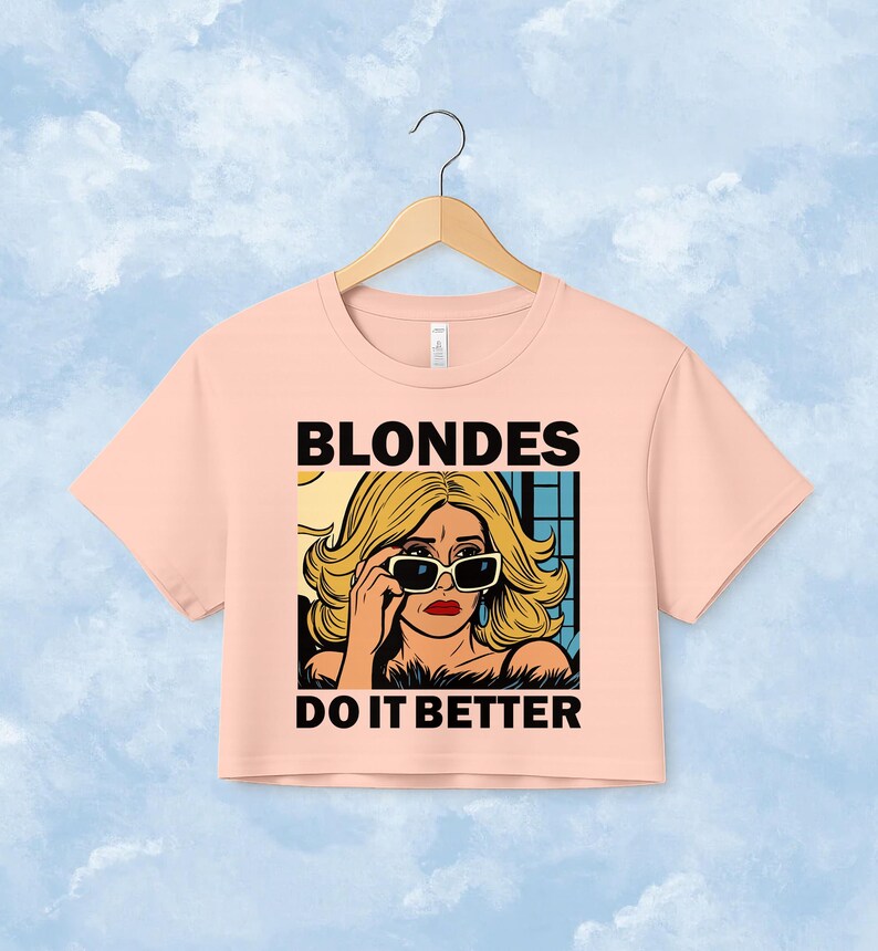 Pu&ograve; includere: Un top corto a maniche corte color pesca chiaro con un'illustrazione grafica di una persona bionda che indossa occhiali da sole. Il testo BLONDES DO IT BETTER &egrave; stampato in grassetto nero in maiuscolo sopra e sotto il disegno centrale.