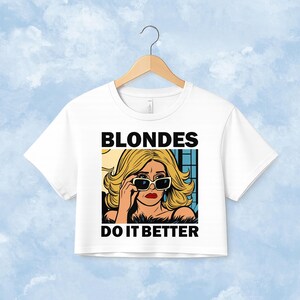 Pu&ograve; includere: Una t-shirt corta bianca con un'illustrazione grafica di una persona bionda che indossa occhiali da sole. Il testo BLONDES DO IT BETTER &egrave; stampato in lettere maiuscole nere sopra e sotto l'illustrazione in stile fumetto.