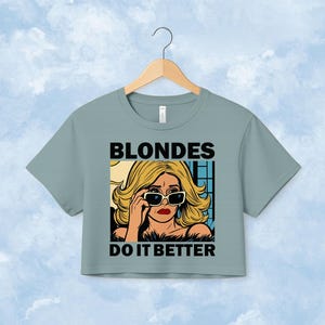 Pu&ograve; includere: Una t-shirt corta color verde salvia con un'illustrazione grafica di una persona bionda che indossa occhiali da sole. Il testo "BLONDES" &egrave; stampato in nero sopra l'illustrazione e il testo "DO IT BETTER" &egrave; stampato in nero sotto di essa.