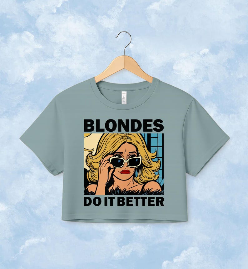 Pu&ograve; includere: Una t-shirt corta color verde salvia con un'illustrazione grafica di una persona bionda che indossa occhiali da sole. Il testo "BLONDES" &egrave; stampato in nero sopra l'illustrazione e il testo "DO IT BETTER" &egrave; stampato in nero sotto di essa.