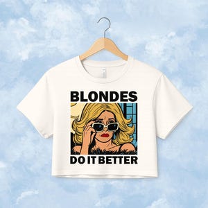Pu&ograve; includere: Un top corto bianco a maniche corte con un'illustrazione grafica di una persona bionda che indossa occhiali da sole. Il testo BLONDES DO IT BETTER &egrave; stampato in grassetto nero in maiuscolo sopra e sotto l'illustrazione in stile fumetto.