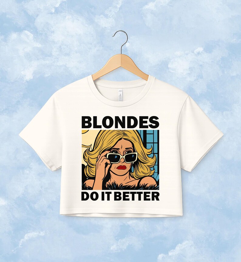 Pu&ograve; includere: Un top corto bianco a maniche corte con un'illustrazione grafica di una persona bionda che indossa occhiali da sole. Il testo BLONDES DO IT BETTER &egrave; stampato in grassetto nero in maiuscolo sopra e sotto l'illustrazione in stile fumetto.