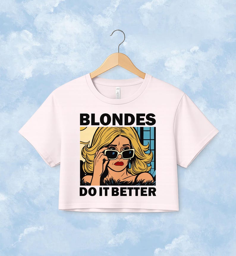 Pu&ograve; includere: Un crop top rosa chiaro con un'illustrazione grafica di una persona bionda che indossa occhiali da sole neri. Il testo "BLONDES" &egrave; stampato in nero sopra l'illustrazione e il testo "DO IT BETTER" &egrave; stampato in nero sotto l'illustrazione.