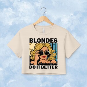 Pu&ograve; includere: Un top corto beige a maniche corte con un'illustrazione grafica di una persona bionda che indossa occhiali da sole. Il testo BLONDES DO IT BETTER &egrave; stampato in grassetto nero in maiuscolo sopra e sotto l'illustrazione in stile fumetto.