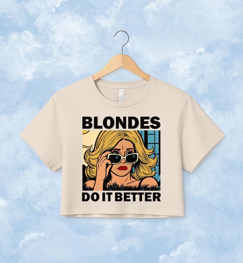 Pu&ograve; includere: Un top corto beige a maniche corte con un'illustrazione grafica di una persona bionda che indossa occhiali da sole. Il testo BLONDES DO IT BETTER &egrave; stampato in grassetto nero in maiuscolo sopra e sotto l'illustrazione in stile fumetto.