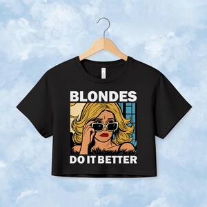 Pu&ograve; includere: Una t-shirt corta nera con una grafica pop art di una persona bionda che indossa occhiali da sole e rossetto rosso. Il testo sopra la grafica recita BLONDES e il testo sotto recita DO IT BETTER in lettere maiuscole bianche e in grassetto.