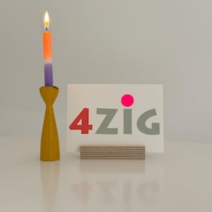 May include: A card with the word "4zig" in gray letters with a pink dot above the "z". The card is sitting on a wooden stand. A candle holder with a lit purple and orange candle is in the background.