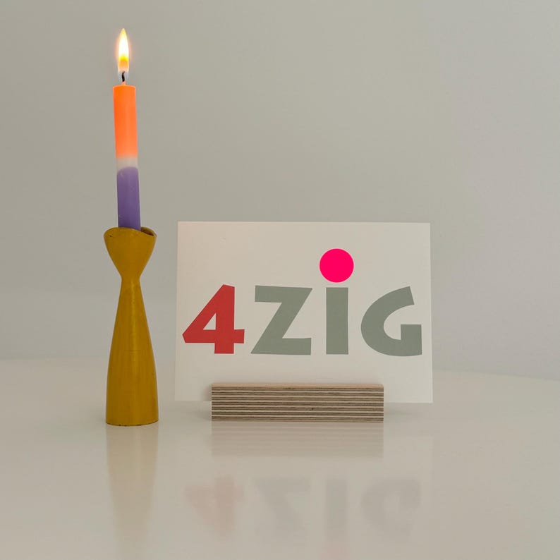 May include: A card with the word "4zig" in gray letters with a pink dot above the "z". The card is sitting on a wooden stand. A candle holder with a lit purple and orange candle is in the background.