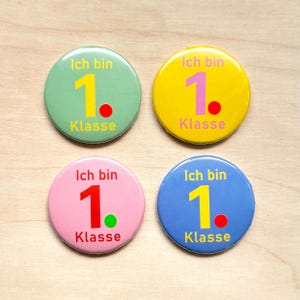 Könnte beinhalten: Vier bunte runde Buttons mit dem deutschen Text "Ich bin 1. Klasse". Die Buttons sind in Grün, Gelb, Rosa und Blau gehalten, jeder mit einer anderen Farbe für die Zahl und den Punkt.