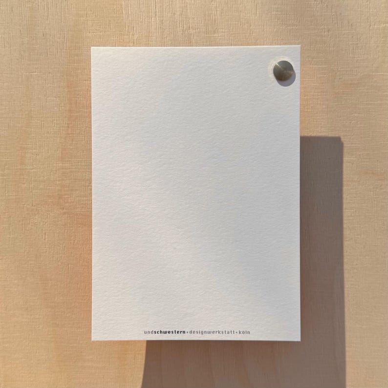 May include: A blank white card with a silver push pin in the top right corner. The card has the text "undschwestern - designwerkstatt - k&ouml;ln" printed in black at the bottom.