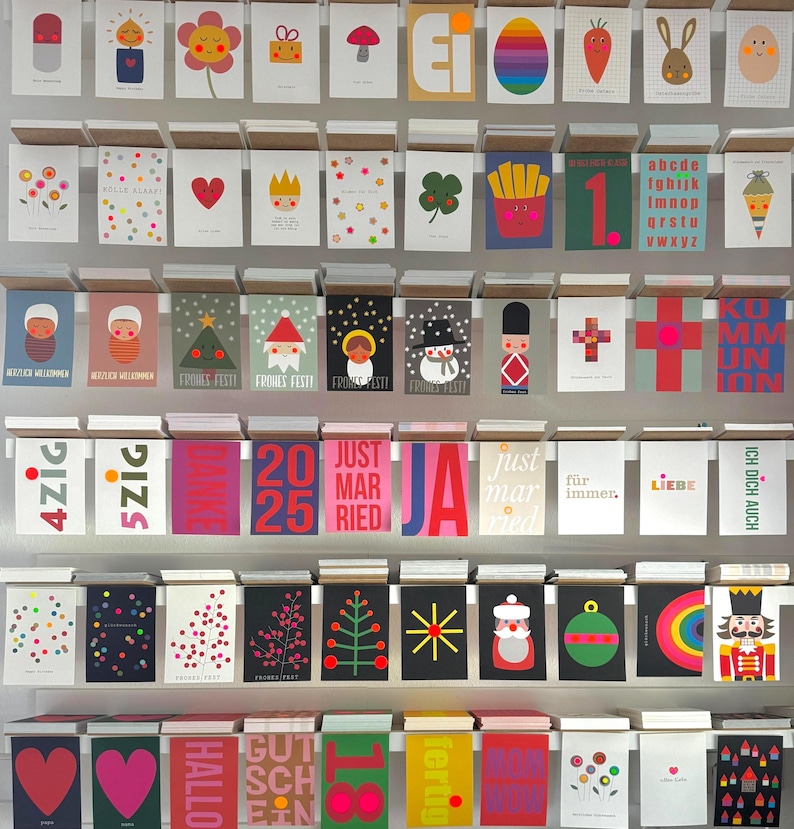 May include: A wall display of colorful greeting cards with various designs and messages. The cards feature illustrations of hearts, flowers, animals, and geometric shapes. Some cards have text, such as "Just Married", "Hallo", and "Ich Dich Auch".