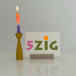 May include: A white card with the text "5zig" in pink, green, and orange letters, sitting on a wooden stand. A yellow candle holder with a purple and orange candle is in the background.