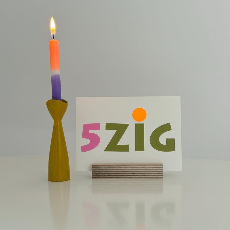 May include: A white card with the text "5zig" in pink, green, and orange letters, sitting on a wooden stand. A yellow candle holder with a purple and orange candle is in the background.