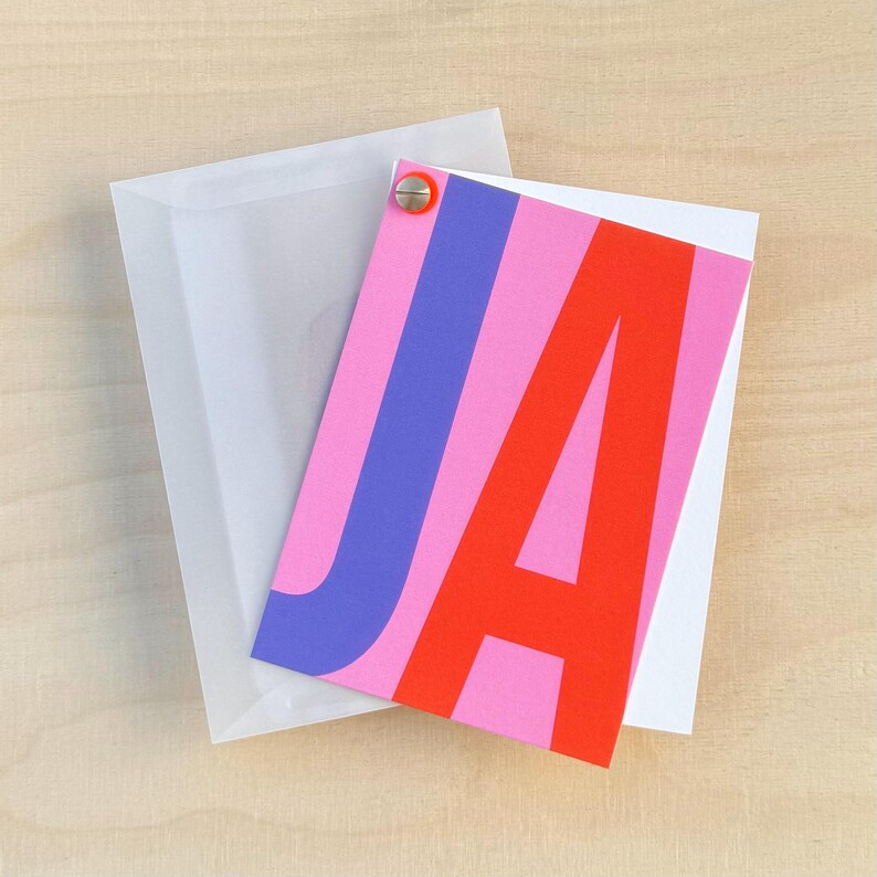 May include: A pink card with the letters "JA" in large, bold, overlapping font. The "J" is blue and the "A" is red. The card is attached to a white envelope with a silver brad.