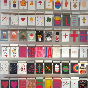 May include: A wall display of colorful greeting cards with various designs and text. The cards feature illustrations of animals, geometric shapes, and holiday themes. Some of the text includes "Just Married", "Frohes Feste", and "Ich Dich Auch".