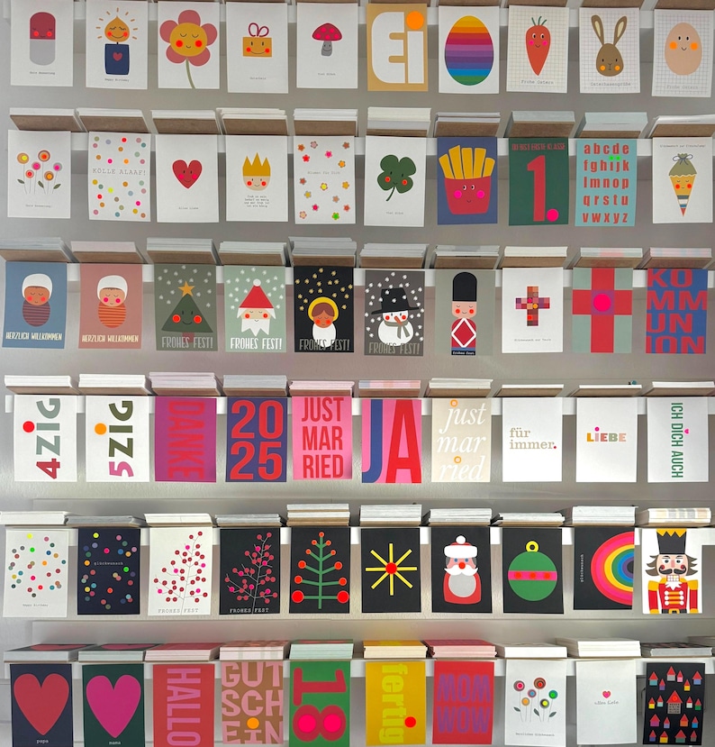 May include: A wall display of colorful greeting cards with various designs and text. The cards feature illustrations of animals, geometric shapes, and holiday themes. Some of the text includes "Just Married", "Frohes Feste", and "Ich Dich Auch".