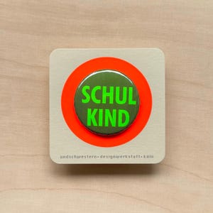 Könnte beinhalten: Ein Button mit den Worten "SCHUL KIND" in grüner Schrift auf dunkelgrünem Hintergrund, umgeben von einem leuchtend orangefarbenen Kreis. Der Button befindet sich auf einer quadratischen, hellbeigen Karte. Der Text "undschwestern designwerkstatt köln" ist unten aufgedruckt.