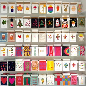 May include: A wall display of colorful greeting cards with various designs and text. The cards are arranged in rows and columns on a white wall. Some of the cards feature illustrations of animals, plants, and geometric shapes. Others have text messages such as "Just Married", "Hallo", and "Pa Pa".