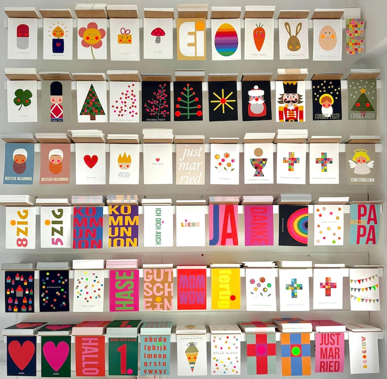 May include: A wall display of colorful greeting cards with various designs and text. The cards are arranged in rows and columns on a white wall. Some of the cards feature illustrations of animals, plants, and geometric shapes. Others have text messages such as "Just Married", "Hallo", and "Pa Pa".