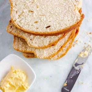 Può includere: Un mucchio di quattro fette di pane bianco, con una consistenza morbida e soffice. Il pane è su una superficie bianca, accanto a una piccola ciotola bianca piena di burro e un coltello da burro argentato.