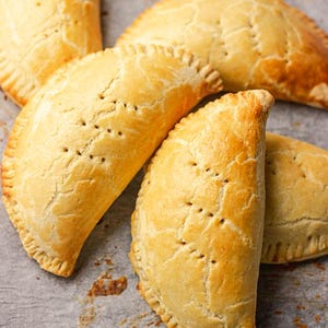 Puede incluir: Cuatro pasteles de color marrón dorado con un borde festoneado, dispuestos en una bandeja para hornear forrada con papel pergamino. Los pasteles tienen una superficie ligeramente texturizada y están ligeramente hinchados.