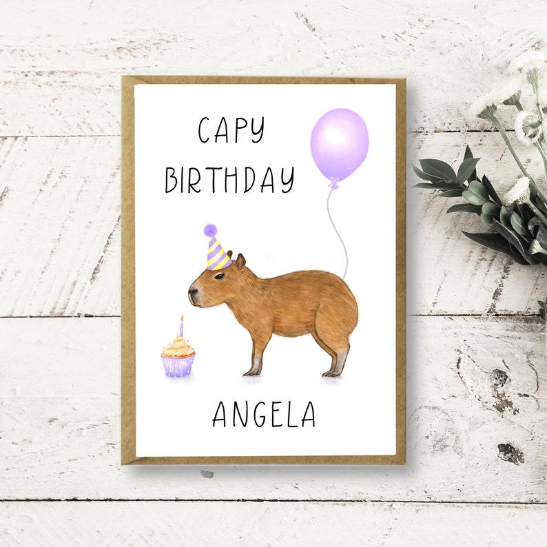 May include: A birthday card featuring a cartoon capybara wearing a party hat and holding a cupcake. The card has the text "Capy Birthday Angela" and a purple balloon.