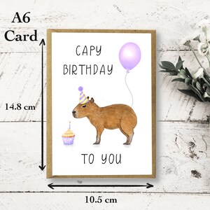 May include: A white card with brown trim, featuring a cartoon capybara wearing a party hat and holding a cupcake. The card reads "Capy Birthday to you" and has a purple balloon with a string attached. The card measures 10.5 cm by 14.8 cm.