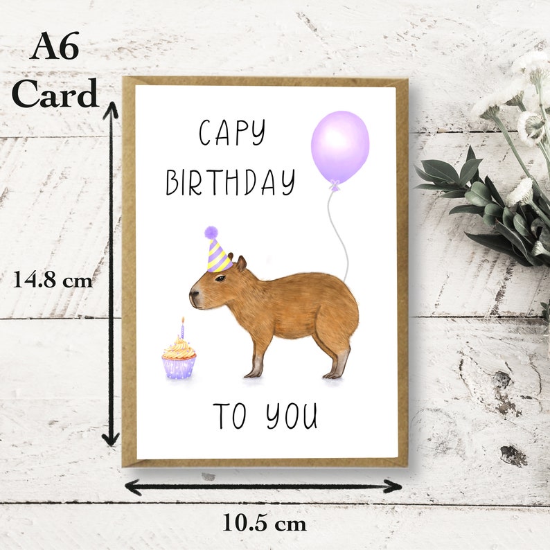 May include: A white card with brown trim, featuring a cartoon capybara wearing a party hat and holding a cupcake. The card reads "Capy Birthday to you" and has a purple balloon with a string attached. The card measures 10.5 cm by 14.8 cm.