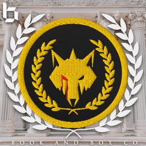 Puede incluir: Un parche bordado amarillo con fondo negro. El parche presenta un lobo amarillo con una lágrima roja, rodeado de una corona de laurel amarilla.