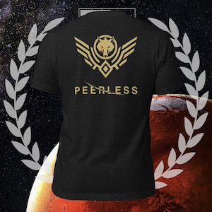 Pode incluir: Camiseta preta com um emblema de lobo dourado e a palavra "PEERLESS" impressa em dourado. O emblema apresenta uma cabeça de lobo com um olho vermelho, asas e formas geométricas. A camiseta é de manga curta.