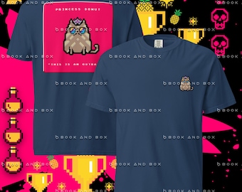 May include: A navy blue t-shirt featuring a pixel art graphic of a cat wearing a crown and blue sunglasses. The back displays a large pink square with the text PRINCESS DONUT and THIS IS AN OUTRAGE, while the front chest shows a small version of the cat.